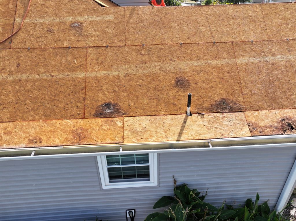 Drone view of bare roof decking after tear-off, showing stained sheathing, and a vent pipe before new underlayment